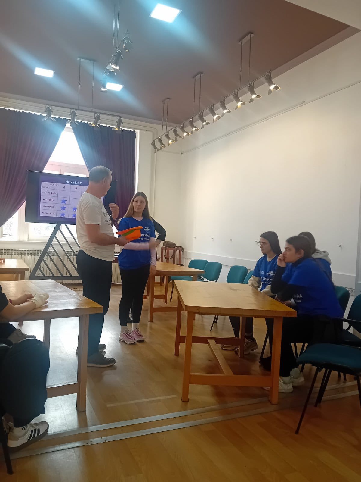 A person standing in a room with people sitting around tables
AI-generated content may be incorrect.